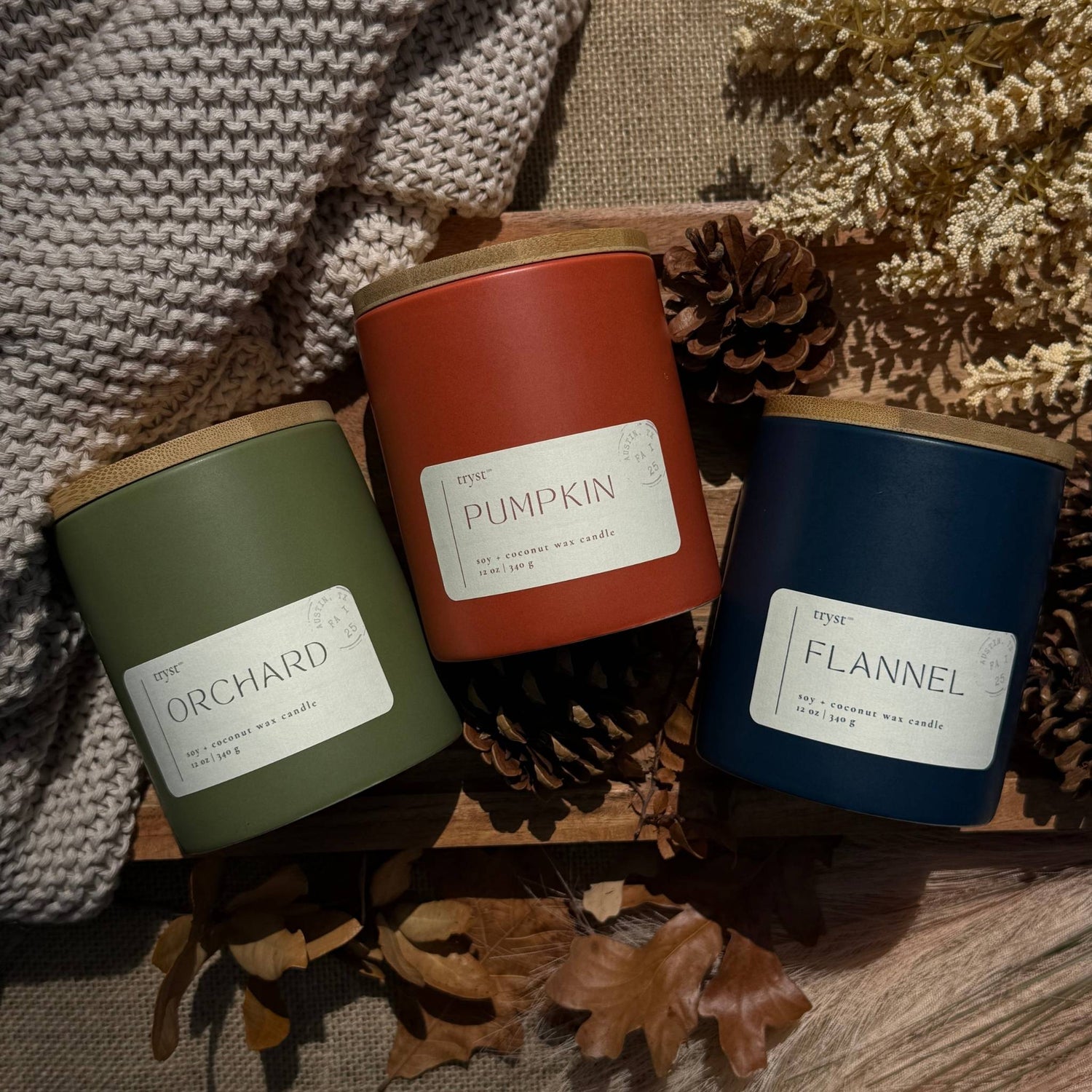 Three scented candles labeled 'Orchard', 'Pumpkin', and 'Flannel' on a textured surface with pinecones and leaves.