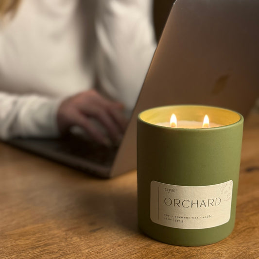 Green candle labeled 'Orchard' on a wooden surface with a blurred person in the background.