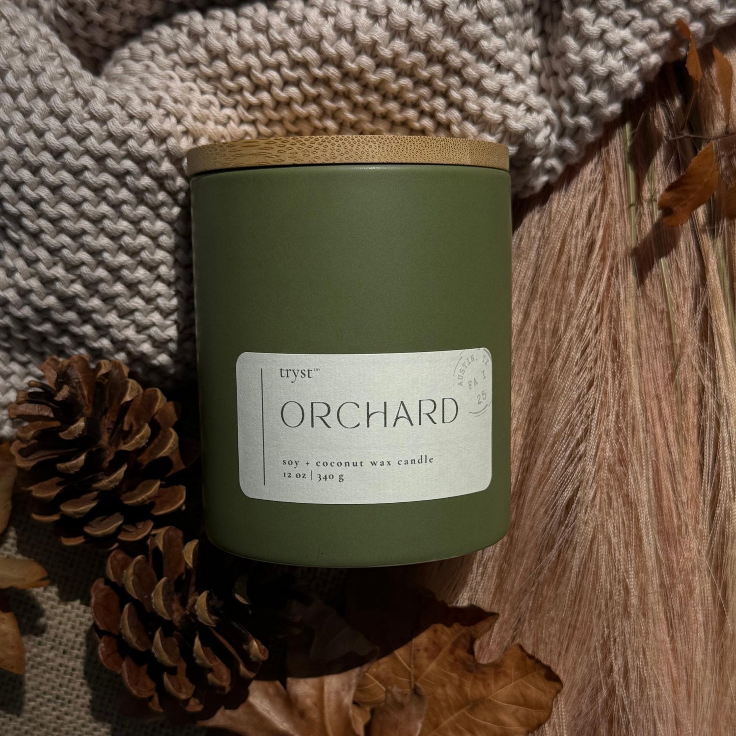 Green candle labeled 'Orchard' with a wooden lid, surrounded by pinecones and leaves on a textured fabric background.