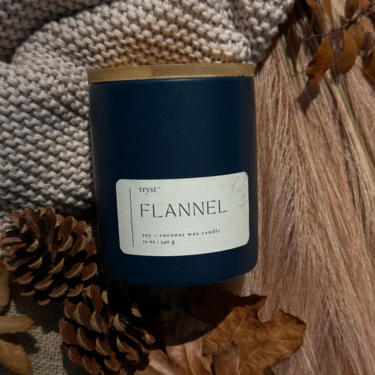 Candle labeled 'Flannel' with a natural setting of leaves and fabric.