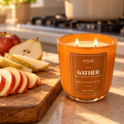 Gather two-wick candle lit in an orange glass jar on a kitchen counter, next to a cutting board of sliced apples and pears