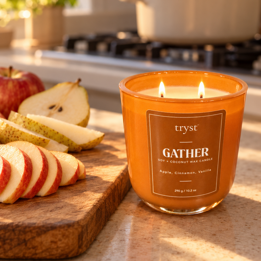 Gather two-wick candle lit in an orange glass jar on a kitchen counter, next to a cutting board of sliced apples and pears