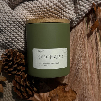 Green candle labeled 'Orchard' with a wooden lid, surrounded by pinecones and leaves on a textured fabric background.