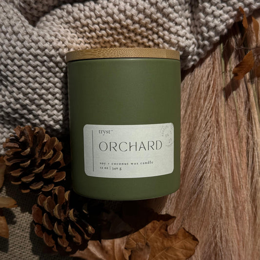 Green candle labeled 'Orchard' with a wooden lid, surrounded by pinecones and leaves on a textured fabric background.
