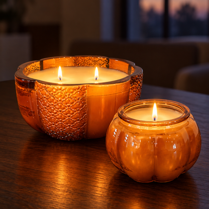 Large two-wick and small one-wick Pumpkin candles lit on a dark wood table at dusk, glowing in amber glass pumpkin jars